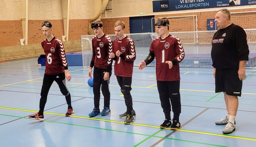 4 unge mænd står på en boldbane klar til at spille. Ved siden af står en træner
