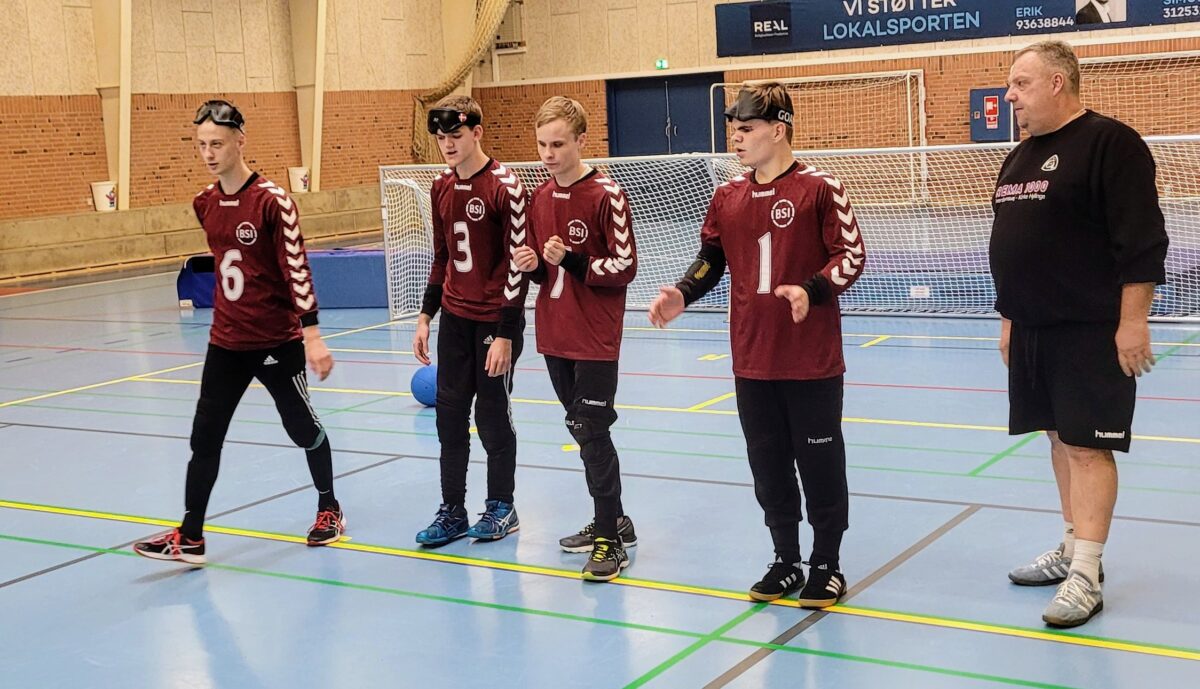 4 unge mænd står på en boldbane klar til at spille. Ved siden af står en træner