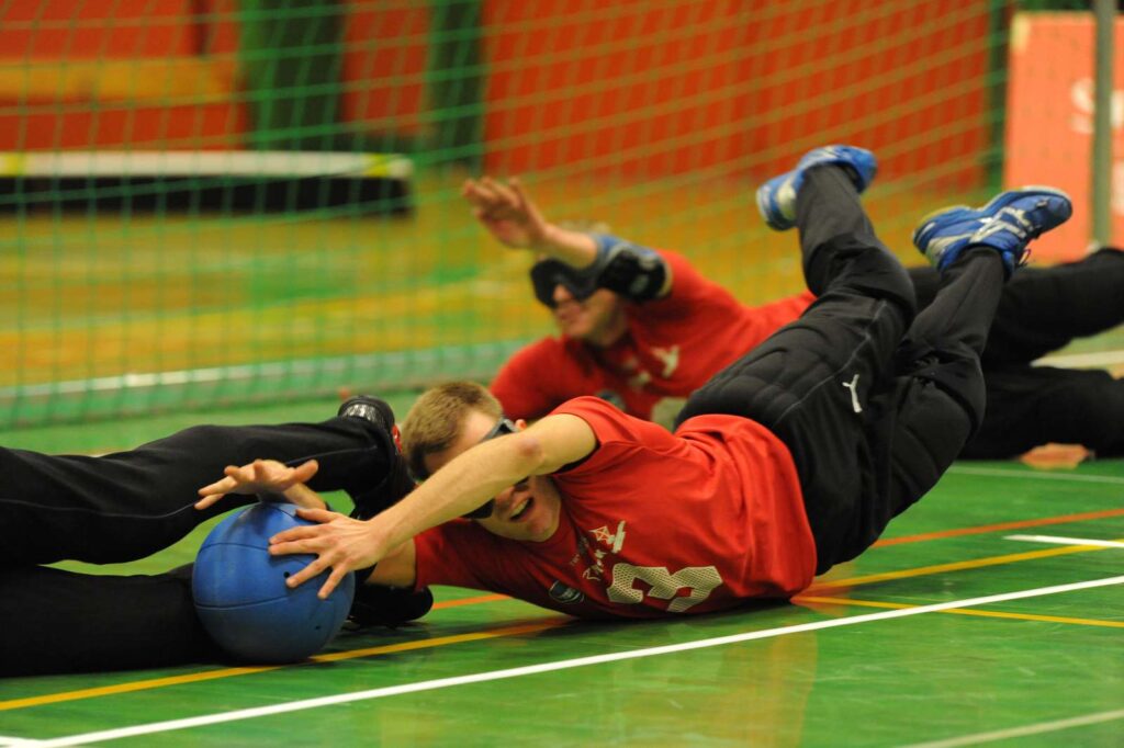 En man ligger på jorden og griber ud efter en bold. Goalballbillede forsvar af herrelandshold af Preben B. Søborg